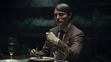 A well-dressed man sits alone at a dimly lit dinner table with a formal place setting, holding a fork and knife, ready to eat. The background is dark and blurred, suggesting an elegant, intimate indoor setting.