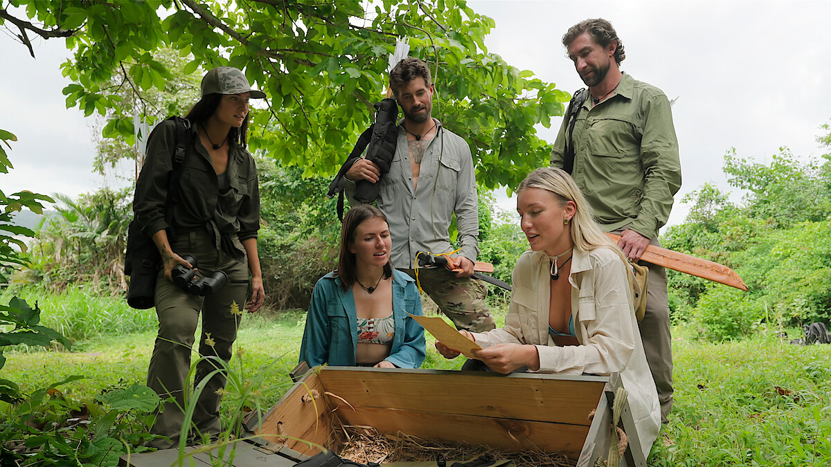 Five people outdoors in a green, forested area gathered around an open wooden chest; one person is holding papers and discussing findings as others watch attentively, with relaxed, adventurous expressions.
