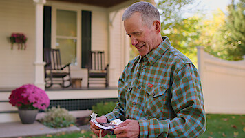 Man in plaid shirt standing in a yard, opening a small package, with a house, porch, rocking chairs, potted flowers, and a white fence in the background on a sunny day.