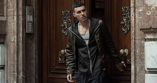 A man wearing a black sweatshirt and brown jacket stands in an open doorway.