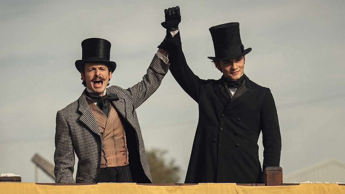 Two men in vintage attire and top hats stand on a platform outdoors, holding hands victoriously, conveying a celebratory and triumphant mood under a cloudy sky, likely at a historical event or reenactment.