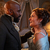 A man and woman in historical costumes gaze at each other affectionately in an ornate room with elegant drapes and soft lighting, suggesting a romantic or intimate moment in a period drama setting.