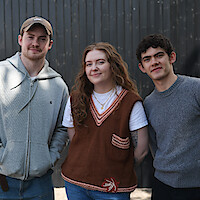 Three young adults standing outside in casual clothing, smiling in front of a dark vertical wooden fence on a bright day.