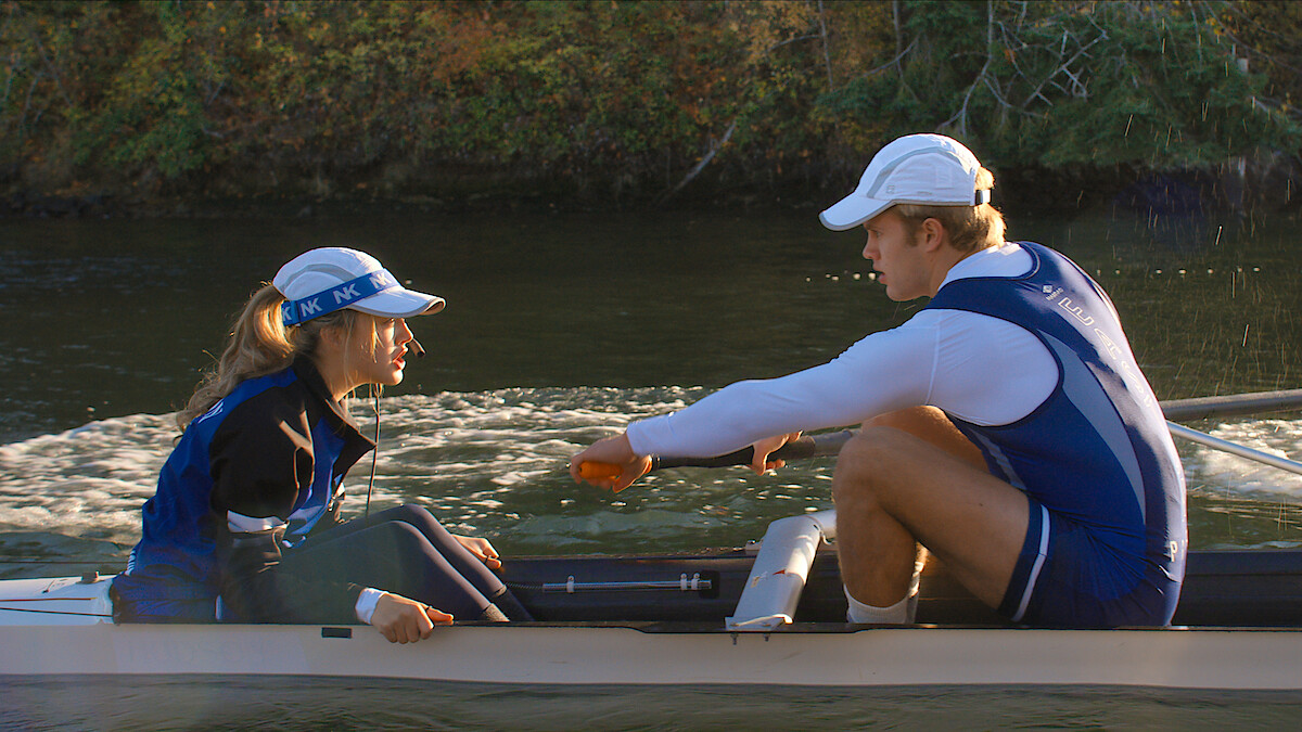 Two rowers in blue uniforms and white caps sit in a racing shell on a calm river, surrounded by greenery, appearing to be in discussion or preparing for a rowing session.