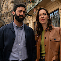 Man and woman standing together outdoors in a city, dressed in smart casual clothing, with historic architecture and building details visible in the background.
