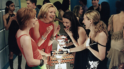 Four women dressed stylishly laugh and enjoy cocktails together around a small table at a lively indoor party with people socializing in the background.
