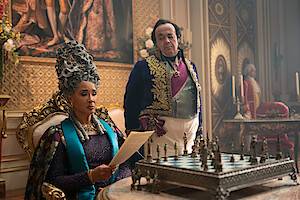 A regal woman in elaborate period attire reads a letter while seated at an ornate chess table in a lavish, opulent room; a man in formal clothing stands behind her, with rich decorations and candelabras in the background.