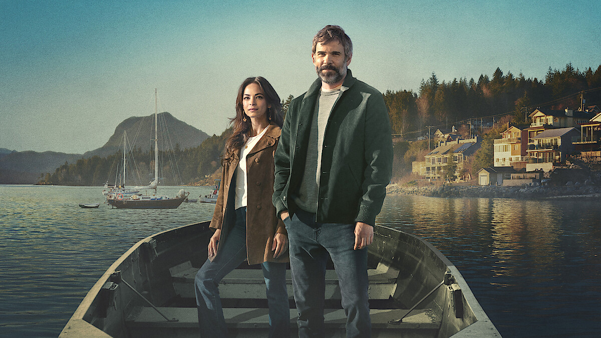 Man and woman standing in a rowboat on calm water, with sailboats, mountains, and a small waterfront town in the background. The setting appears peaceful, natural, and scenic.
