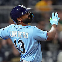 Baseball player in blue Tampa Bay Rays uniform and helmet mid-swing during a game, with stadium crowd and lights in the background. Action shot captures motion and intensity of the sport.