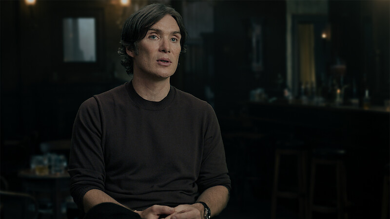 A man with short dark hair sits and speaks in a dimly lit bar or pub setting, with tables, chairs, and a bar counter visible in the background. The atmosphere is moody and subdued.