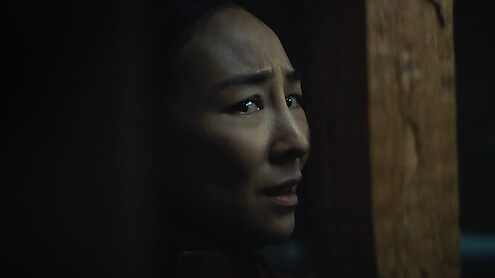 Worried woman in dim lighting, partially hidden behind a wooden beam, looking over her shoulder in a tense, shadowy environment.