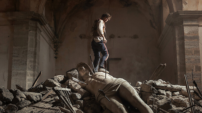 A person stands on rubble inside a damaged, old stone building, with a large broken statue lying among the debris, creating a dramatic and somber atmosphere.