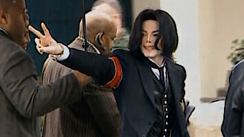 Person in a black suit with red armband making a peace sign outdoors, surrounded by several people in a public or official setting.