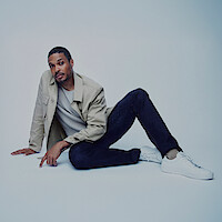 Ray Fisher wears dark jeans and a tan jacket, and sits in a jaunty pose.