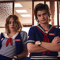 Two young adults in sailor-themed uniforms stand in a brightly lit room with glass doors, looking directly at the camera. The mood is casual and slightly serious.