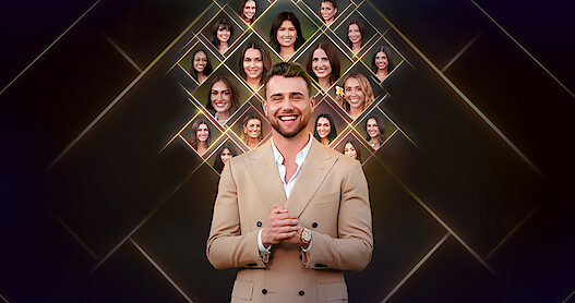 A smiling man in a tan suit stands in front of a dark background with glowing diamond shapes featuring multiple women’s faces displayed above and behind him, suggesting a reality TV or dating show setting.