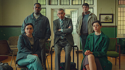 Five adults, three seated and two standing, pose in a serious group setting inside a classroom or office with green walls, wooden floor, chairs, and a painting on the wall, suggesting a meeting or discussion environment.