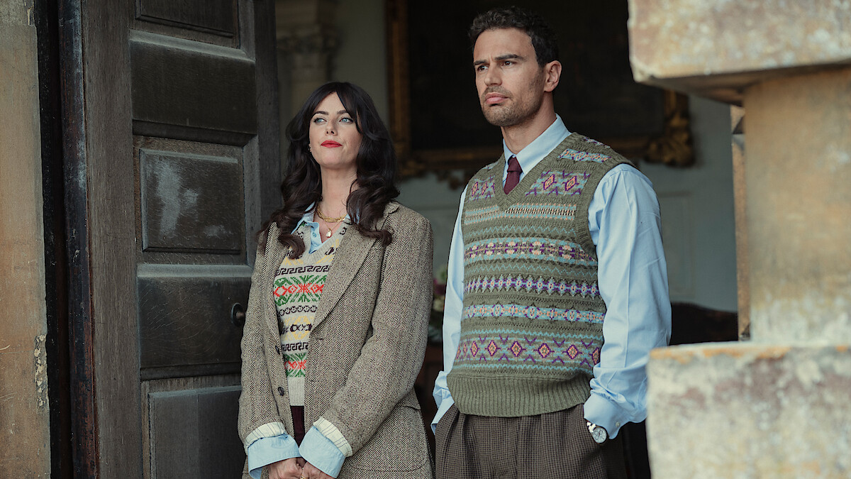 A woman and a man in vintage sweater vests and formal clothing stand side by side in front of a large open door, with stone architecture visible, looking thoughtfully into the distance.