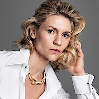 Woman with blonde wavy hair wearing a white shirt and gold necklace, seated against a plain gray background, posing thoughtfully with her hand touching her head, creating a calm and introspective mood.
