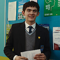 A smiling student in school uniform stands indoors holding papers, with a blue bulletin board behind featuring posters about mental health and financial literacy.