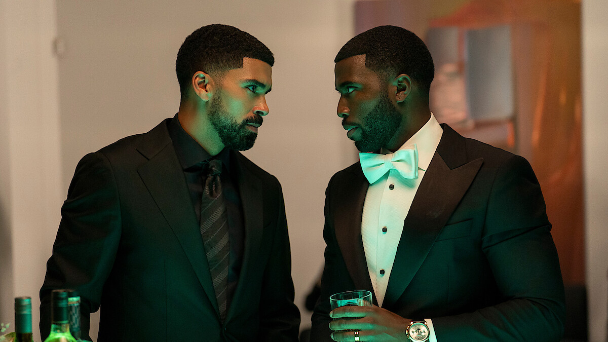 Two men in formal black suits stand close, engaged in a serious conversation at an indoor event. One holds a drink, bottles are visible on the table, and ambient green lighting enhances the elegant, upscale setting.
