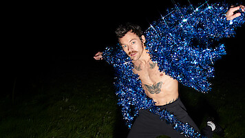 Man with mustache posing outdoors at night, wearing a sparkling blue tinsel jacket open to show his chest and tattoos, standing on grass with a dark background, in a dramatic and playful stance.