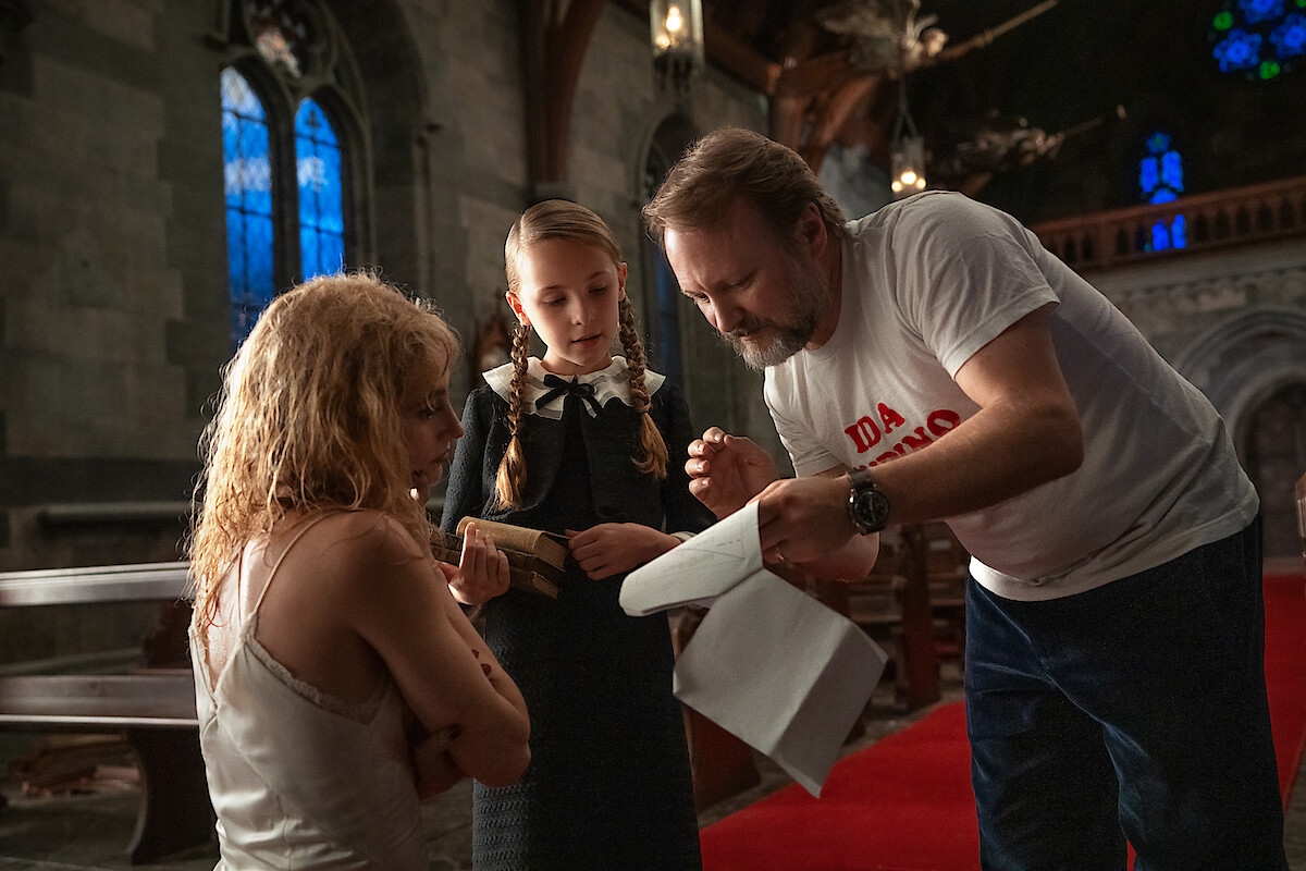 A man gives instructions to two women, one seated and distraught, the other in a school uniform, inside a dimly lit church with stained glass windows, wooden pews, and a serious, contemplative atmosphere.