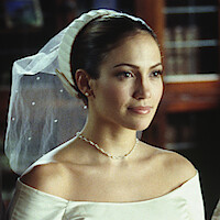 A woman in a white off-shoulder wedding dress with a veil and pearl necklace, standing indoors with a soft background, possibly a library or study with wooden shelves and books.