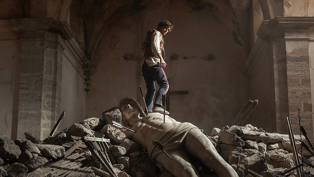 A person stands on rubble inside a damaged, old stone building, with a large broken statue lying among the debris, creating a dramatic and somber atmosphere.