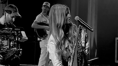 Woman singing into a microphone on stage with a camera crew filming and a musician playing guitar in the background during a live performance or rehearsal, in a studio setting.