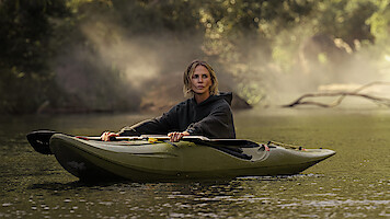 A person wearing a dark hoodie is paddling a green kayak on a calm river, surrounded by trees with mist rising in the background, creating a serene outdoor nature scene.