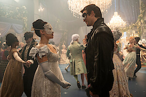 Two people in elegant masquerade costumes face each other at a lavish masked ball, surrounded by dancers in formal attire and ornate masks, in a grand, chandelier-lit ballroom decorated with drapery and greenery.