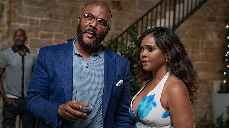 A man in a blue blazer holding a wine glass stands next to a woman in a floral dress at an upscale indoor gathering with stone walls, greenery, and a staircase in the background. Another person is blurred in the distance.