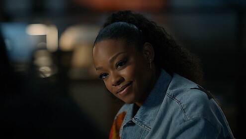 A woman with curly hair tied back is sitting indoors, wearing a denim jacket, and smiling warmly in a softly lit environment with a blurred background.