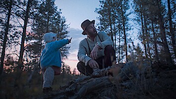 A man in a hat and a child in a blue sweater explore a forest at dusk, surrounded by tall trees. The child points at something, creating a sense of curiosity and peacefulness in the natural setting.