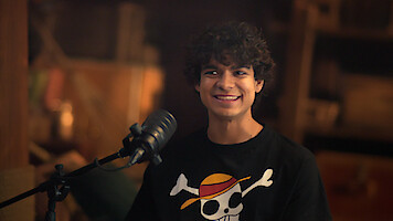 Iñaki Godoy sits indoors in a warmly lit, cozy room, smiling as he faces a microphone. He wears a black shirt featuring a skull with a straw hat design. Behind him, wooden shelves and soft décor add to the inviting, relaxed atmosphere.