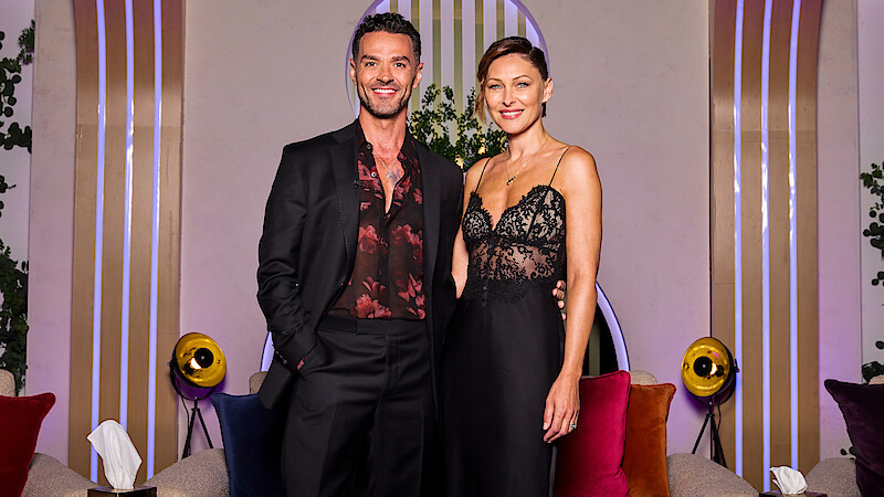 A man in a black suit and a woman in a black lace dress stand together smiling in a modern lounge with colorful cushions, round lights, plants, and decorative vertical lines in the background.