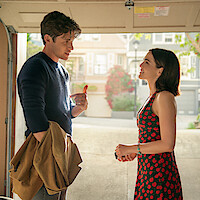 A man holding a coat talks to a smiling woman in a cherry dress inside a house garage with sunlight coming in, suburban homes and greenery visible outside in the background.
