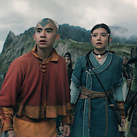 Two young people in fantasy clothing stand side by side, looking ahead with concern. They are outside in a mountainous landscape with cloudy skies and others visible in the background.
