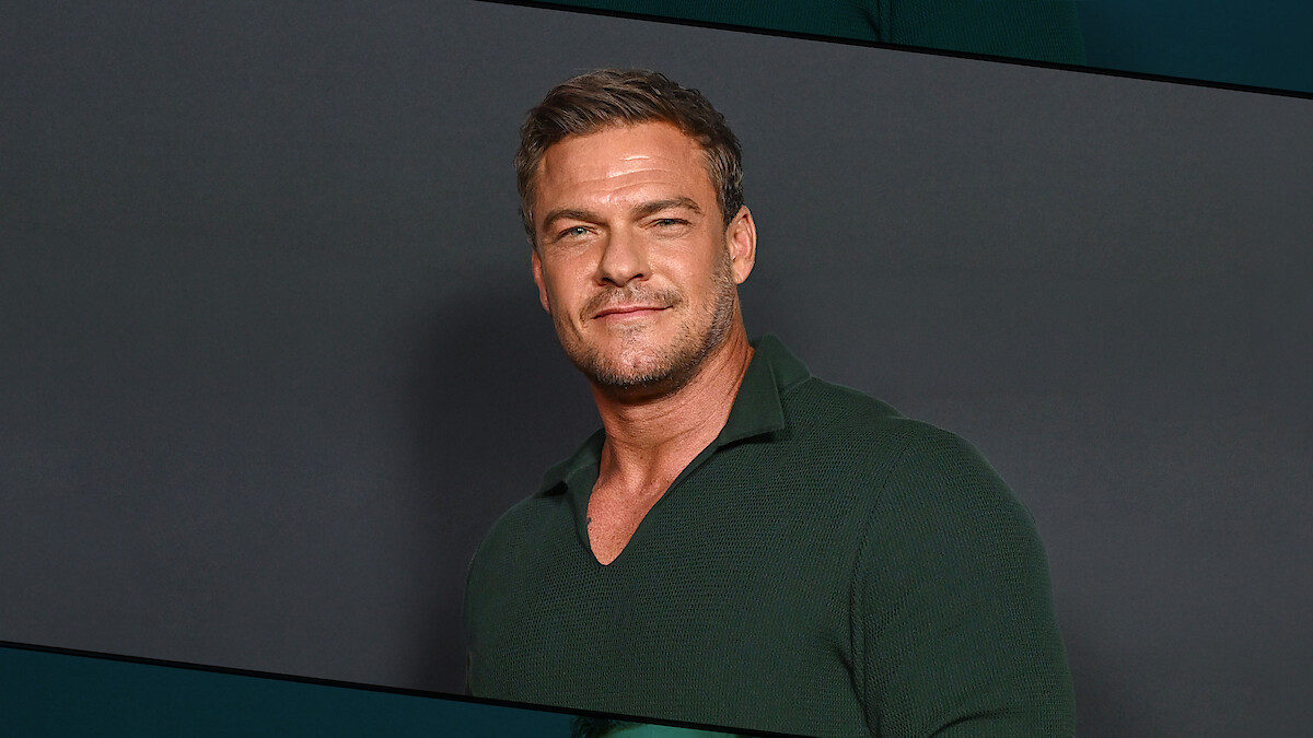 Alan Ritchson with short hair wearing a dark green collared shirt standing against a plain dark background, facing forward and smiling slightly in a studio setting.