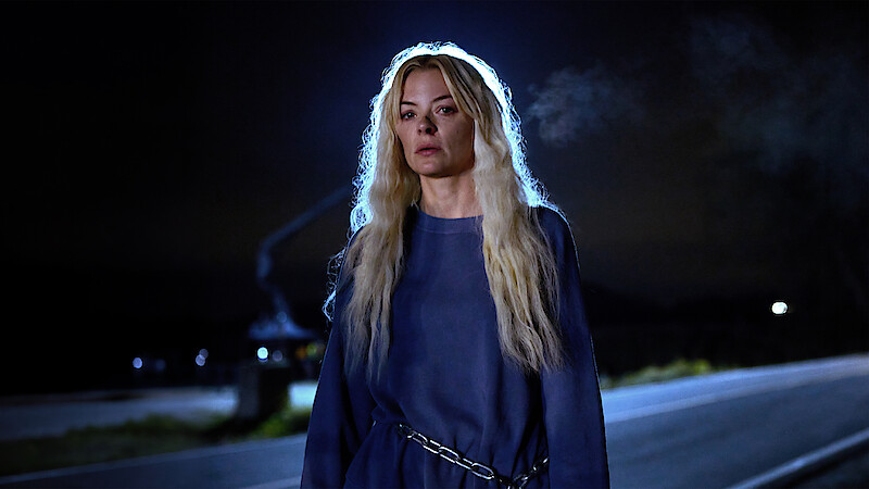 A woman with long blonde hair stands alone on a dark road at night, wearing a blue outfit and chains around her waist. Her breath is visible in the cold air, and the scene is illuminated by a faint blue backlight.