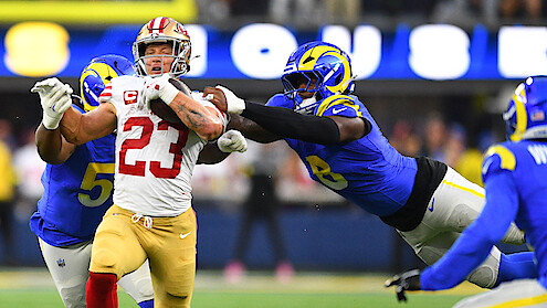 Football player in red and gold uniform running with the ball, being tackled by two players in blue and yellow uniforms on a professional field during a game. Stadium lights and blurred audience in the background.