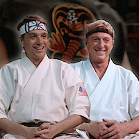 Ralph Macchio and William Zabka sit together in karate uniforms smiling looking at a young Ralph Macchio in 'The Karate Kid.'