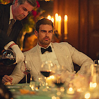Man in white tuxedo sits at elegant candlelit dinner table as a waiter pours red wine into his glass, surrounded by flowers, glassware, and formal dining settings in a luxurious, warmly lit room.