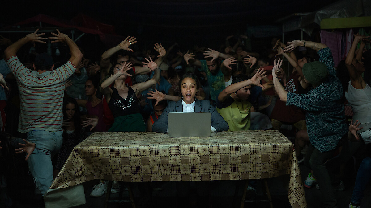 Rita (Zoe Saldaña) sits at a table behind a laptop, as people behind her dance wildly.