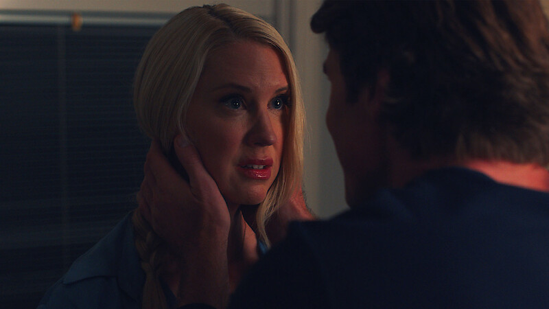 A blonde woman looks anxious as a man gently holds her face in a dimly lit room, suggesting a tense or emotional moment between the two people indoors at night.
