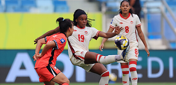 Female soccer players in action during a match, one in a white uniform controls the ball with her foot as opponents and teammates watch, in a stadium with blue seats and a colorful background.
