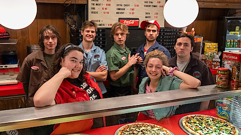 Seven young people in pizza shop uniforms stand behind a glass counter with pizzas, smiling and posing for a photo; wood paneling, a menu board, and snack shelves are visible in the cozy, retro-styled restaurant environment.