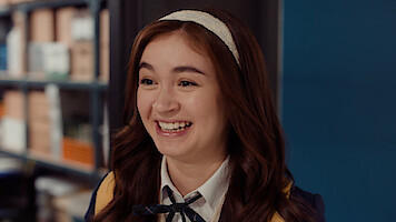 Smiling young woman with a headband and school uniform in an indoor setting, standing near blue wall and bookshelves.