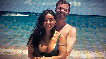 A smiling couple standing together on a sunny beach with waves and a blue ocean in the background.